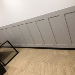 USŁUGI BUDOWLANO-REMONTOWE GRZEGORZ PAŹDZIORA - Szara boazeria angielska na skośnej ścianie, podłoga z paneli ułożonych w jodełkę, stolik kawowy z czarnymi nogami i drewnianym blatem.