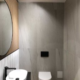 USŁUGI BUDOWLANO-REMONTOWE GRZEGORZ PAŹDZIORA - Nowoczesna łazienka w odcieniach szarości z drewnianymi akcentami, owalne lustro z podświetleniem, minimalistyczna umywalka nablatowa z czarną armaturą, podwieszana toaleta i płytki imitujące marmur.