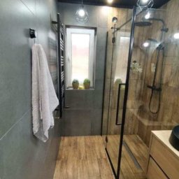 USŁUGI BUDOWLANO-REMONTOWE GRZEGORZ PAŹDZIORA - Nowoczesna łazienka z szarymi płytkami ściennymi, drewnopodobną podłogą, kabiną prysznicową typu walk-in z czarnymi ramami, grzejnikiem drabinkowym i oknem z roślinami doniczkowymi.