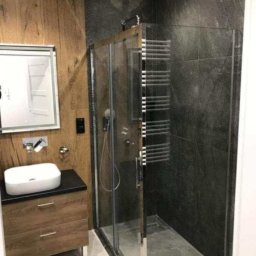 USŁUGI BUDOWLANO-REMONTOWE GRZEGORZ PAŹDZIORA - Nowoczesna łazienka z kabiną prysznicową z chromowanymi detalami, umywalką nablatową na drewnianej szafce i ścianami wykończonymi w drewnie i ciemnym kamieniu.
