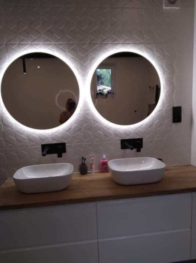 Nowoczesna łazienka z dwoma okrągłymi lustrami z podświetleniem LED, umywalkami nablatowymi na drewnianym blacie i białymi szafkami, ściana wyłożona białą trójwymiarową mozaiką.