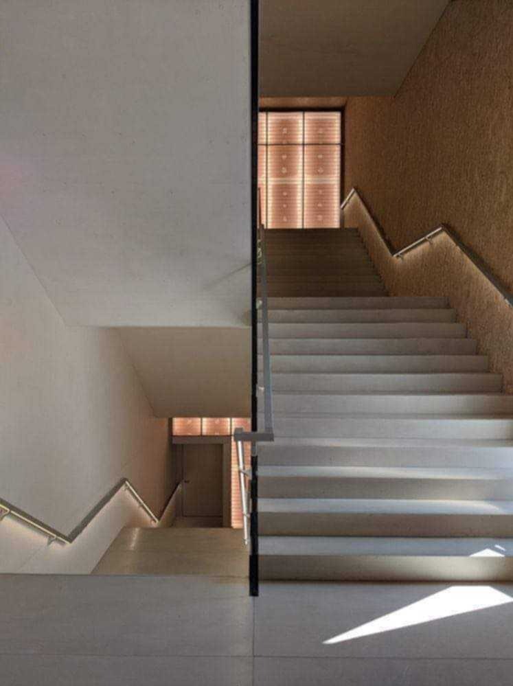 Nowoczesne schody z podświetlanymi poręczami, prowadzące na dwa poziomy, z widocznym fragmentem ściany o fakturze baranka i geometrycznym oświetleniem w tle.