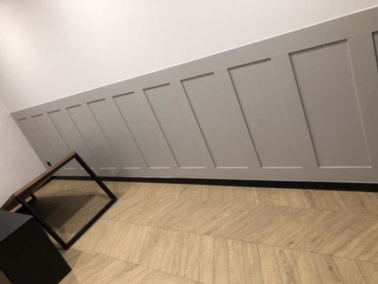 Szara boazeria angielska na skośnej ścianie, podłoga z paneli ułożonych w jodełkę, stolik kawowy z czarnymi nogami i drewnianym blatem.