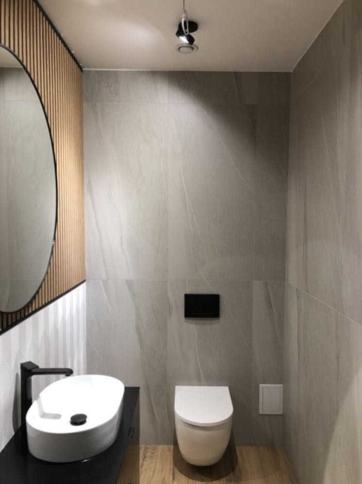 Nowoczesna łazienka w odcieniach szarości z drewnianymi akcentami, owalne lustro z podświetleniem, minimalistyczna umywalka nablatowa z czarną armaturą, podwieszana toaleta i płytki imitujące marmur.