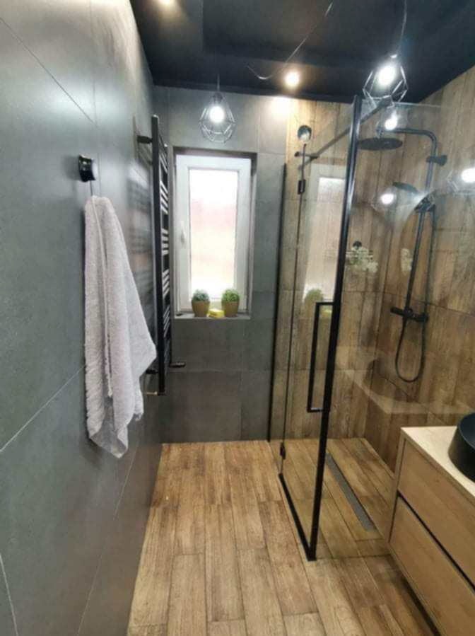 Nowoczesna łazienka z szarymi płytkami ściennymi, drewnopodobną podłogą, kabiną prysznicową typu walk-in z czarnymi ramami, grzejnikiem drabinkowym i oknem z roślinami doniczkowymi.
