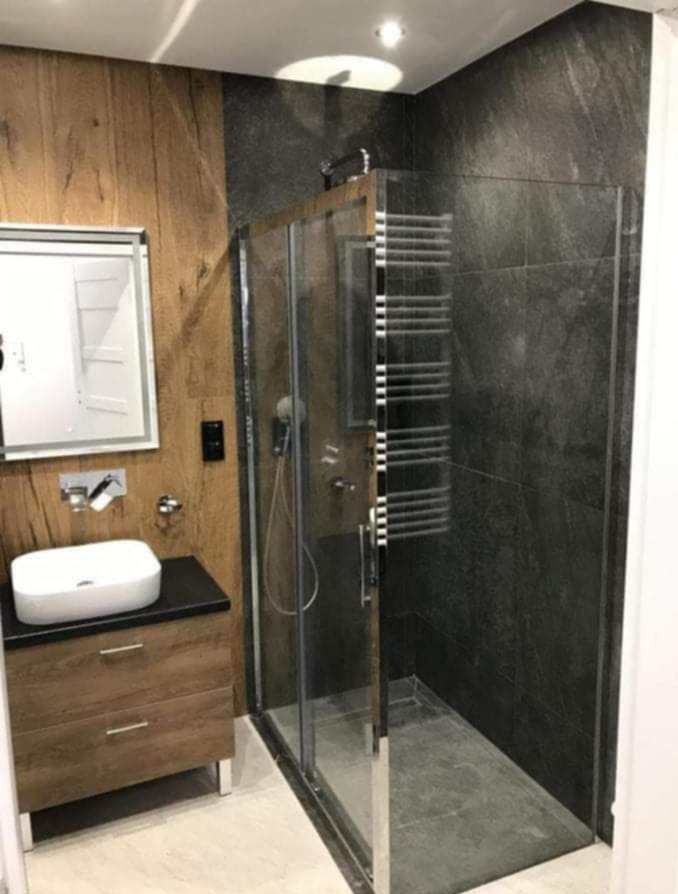 Nowoczesna łazienka z kabiną prysznicową z chromowanymi detalami, umywalką nablatową na drewnianej szafce i ścianami wykończonymi w drewnie i ciemnym kamieniu.
