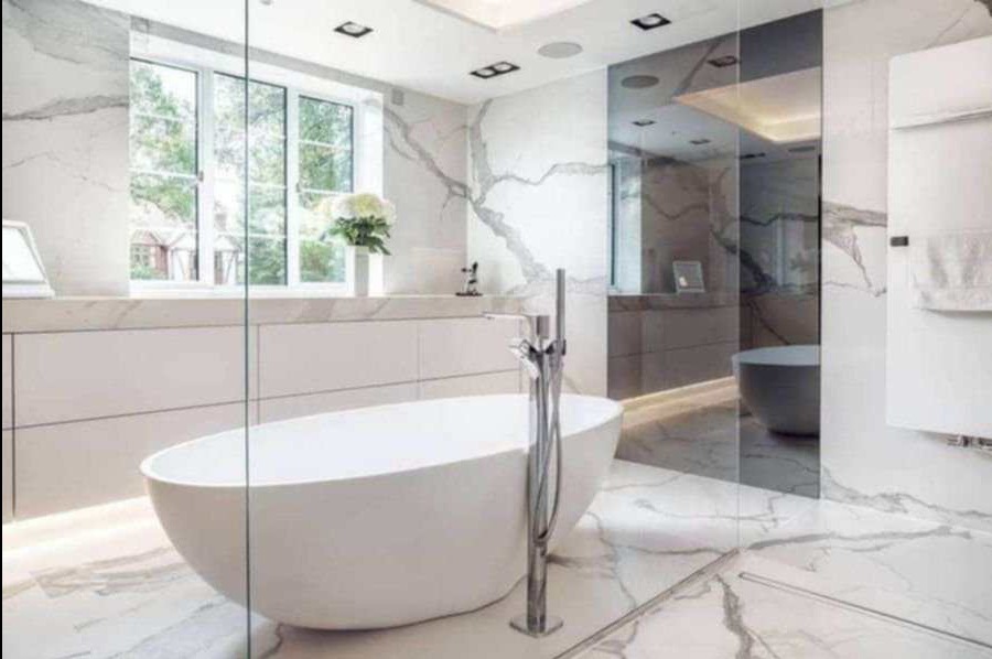Nowoczesna łazienka z wolnostojącą wanną, marmurowymi płytami na ścianach i podłodze, przeszklonym prysznicem i dużym oknem z widokiem na zieleń.