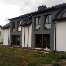 Hubert Szczepaniak - Dwukondygnacyjny dom w trakcie wykańczania elewacji, z szarymi i białymi fragmentami ścian, brązowymi oknami i drzwiami, ciemną dachówką i widoczną osobą pracującą przy ziemi.