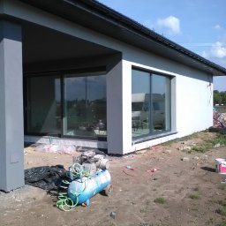 Hubert Szczepaniak - Świeżo otynkowany budynek z dużymi oknami przesuwnymi, widoczny kompresor i akcesoria na placu budowy.