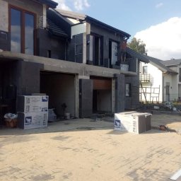 Hubert Szczepaniak - Budynek w trakcie ocieplania elewacji zewnętrznej styropianem grafitowym, widoczny pracownik na wysokości, paczki styropianu na podłożu, dom w stanie surowym otwartym.