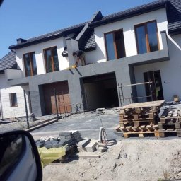 Hubert Szczepaniak - Dwukondygnacyjny dom w trakcie prac wykończeniowych elewacji, widoczny pracownik na balkonie, ułożona kostka brukowa i palety na podjeździe.