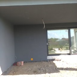 Hubert Szczepaniak - Szara elewacja domu jednorodzinnego po tynkowaniu, widoczne okno przesuwne i przygotowany teren pod wylewkę.