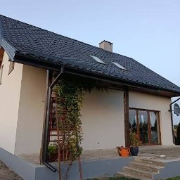 Hubert Szczepaniak - Jasny dom jednorodzinny z ciemnym dachem i oknami dachowymi, widok z boku na elewację z elementami zieleni pnącej, doniczkami i schodami.