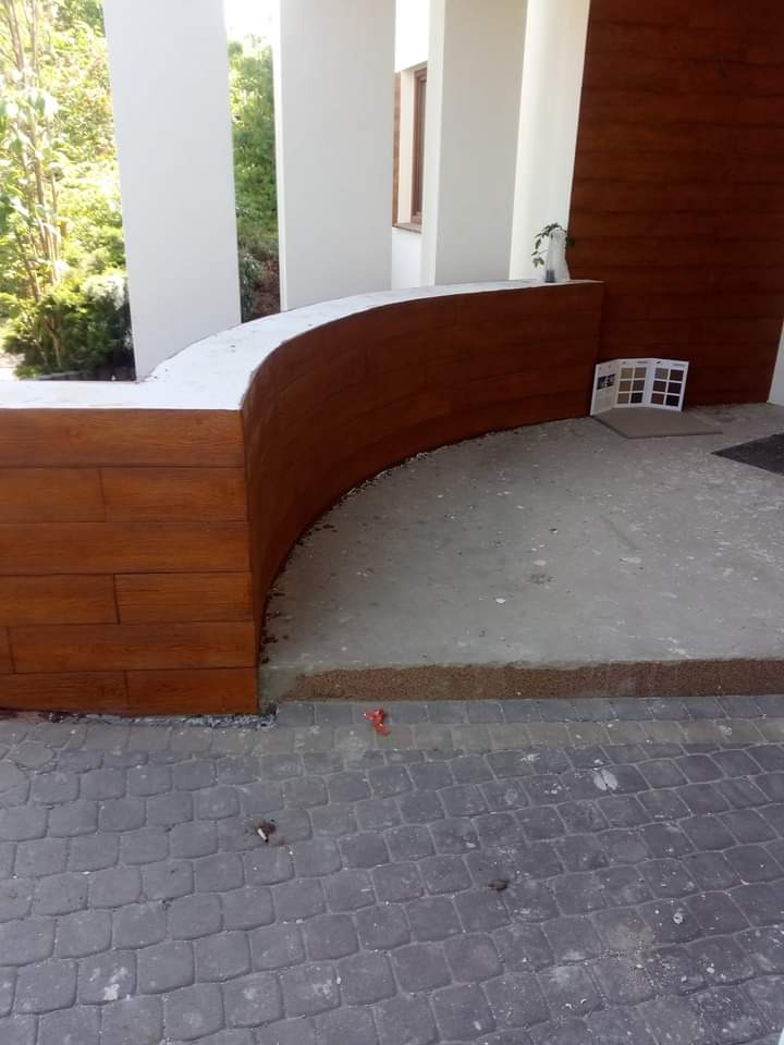 Kątowe ujęcie fragmentu elewacji z drewnianą okładziną i białymi kolumnami, z katalogiem kolorów na podłodze i kostką brukową na pierwszym planie.