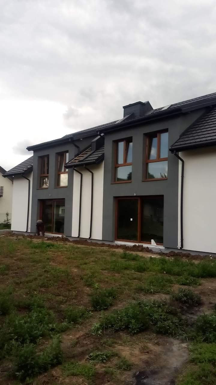 Dwukondygnacyjny dom w trakcie wykańczania elewacji, z szarymi i białymi fragmentami ścian, brązowymi oknami i drzwiami, ciemną dachówką i widoczną osobą pracującą przy ziemi.