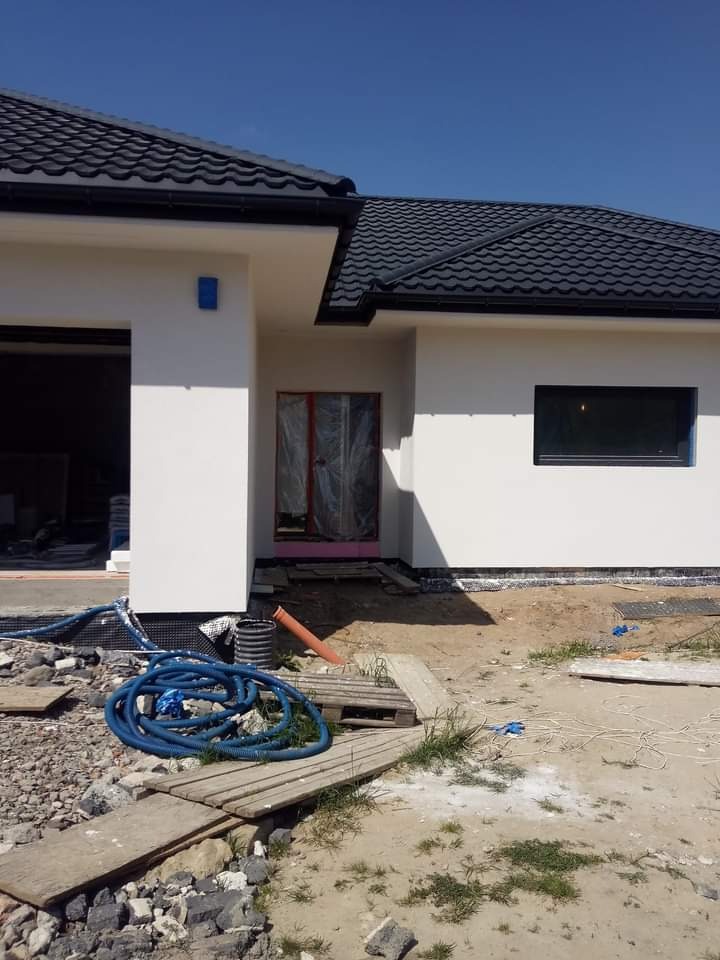 Nowoczesny dom jednorodzinny z białą elewacją i czarnym dachem w trakcie prac wykończeniowych. Widoczne zabezpieczone folią drzwi wejściowe, okno, niebieski wąż ogrodowy i tymczasowe przejście...