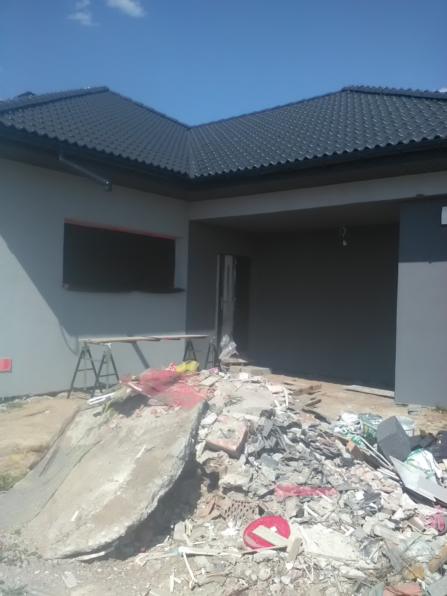 Dom jednorodzinny w trakcie prac wykończeniowych elewacji, z widocznym stertą gruzu przed budynkiem, zabezpieczonym oknem i rusztowaniem.