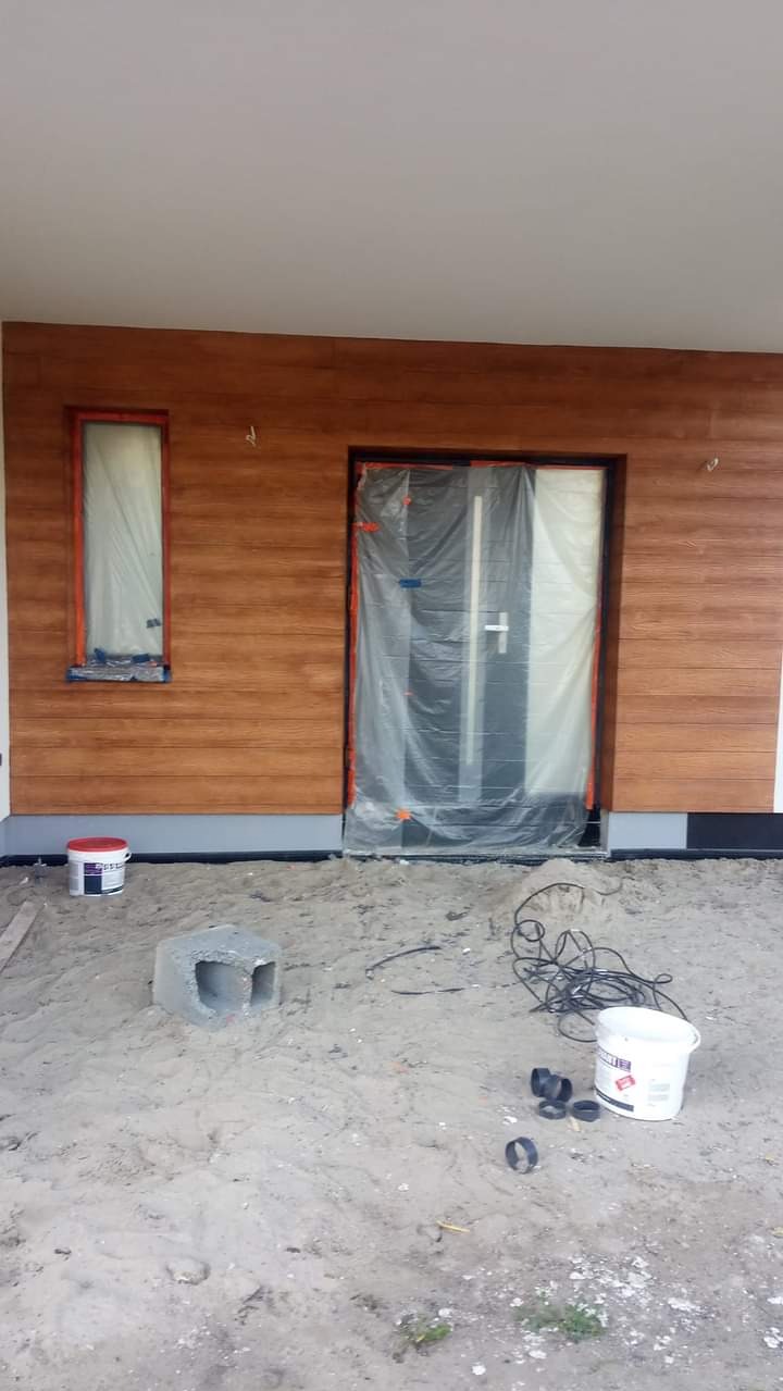 Elewacja domu z drewnopodobną okładziną, okno i drzwi zabezpieczone folią malarską, wiadro farby i betonowy bloczek na piaszczystym podłożu.