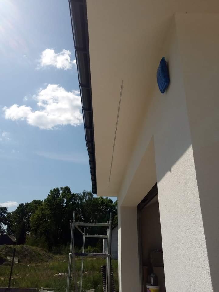 Fragment elewacji budynku z widocznym rusztowaniem, zabezpieczonym elementem w niebieskiej folii i świeżo nałożonym jasnym tynkiem strukturalnym, sfotografowany w słoneczny dzień.