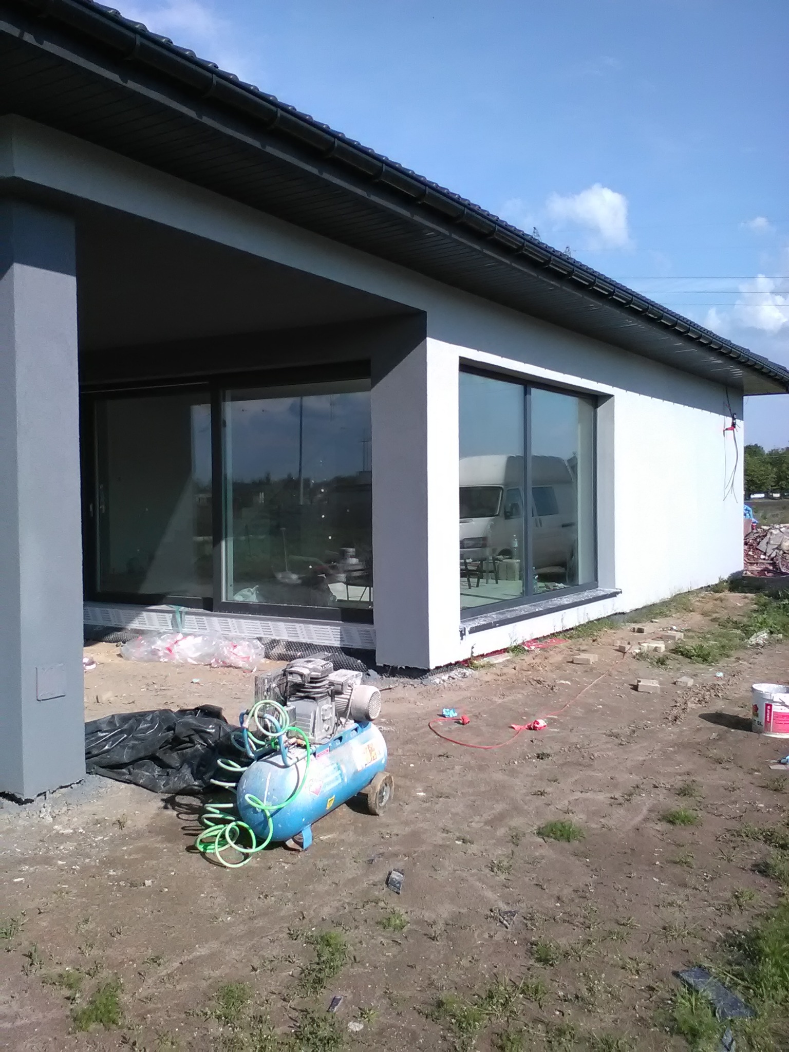 Świeżo otynkowany budynek z dużymi oknami przesuwnymi, widoczny kompresor i akcesoria na placu budowy.