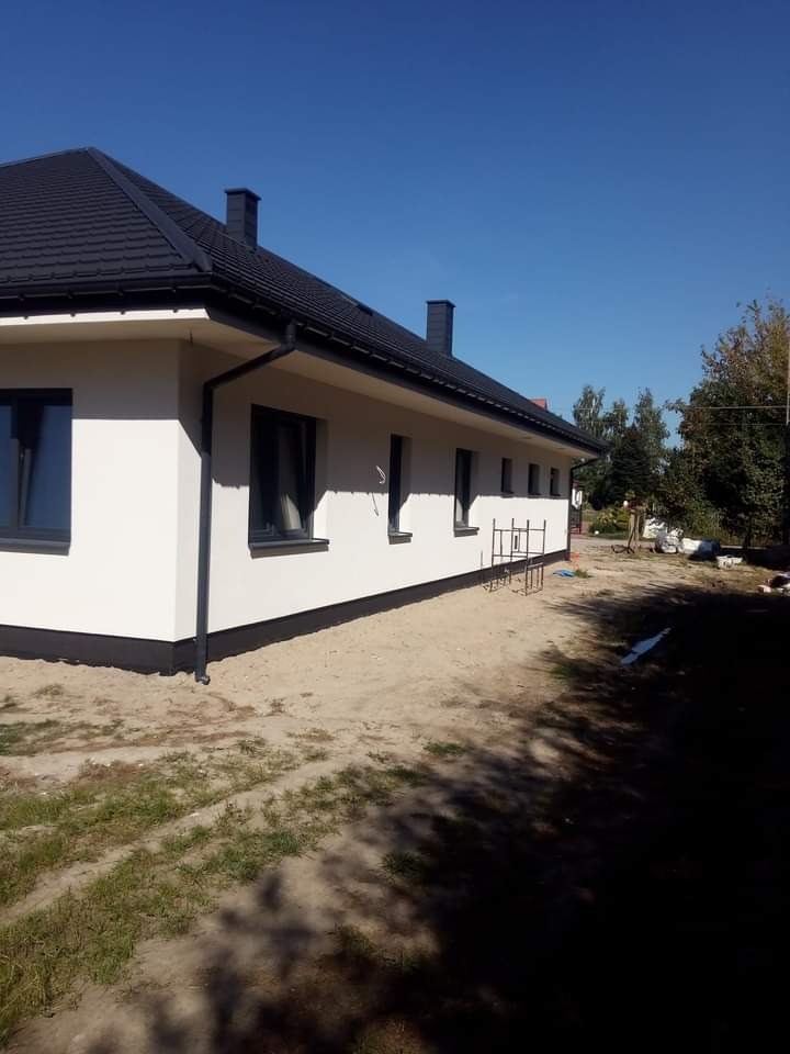 Świeżo pomalowana elewacja domu jednorodzinnego w Żyrardowie, z widocznymi czarnymi detalami wykończeniowymi, takimi jak rynny i obramowania okien, na tle błękitnego nieba.