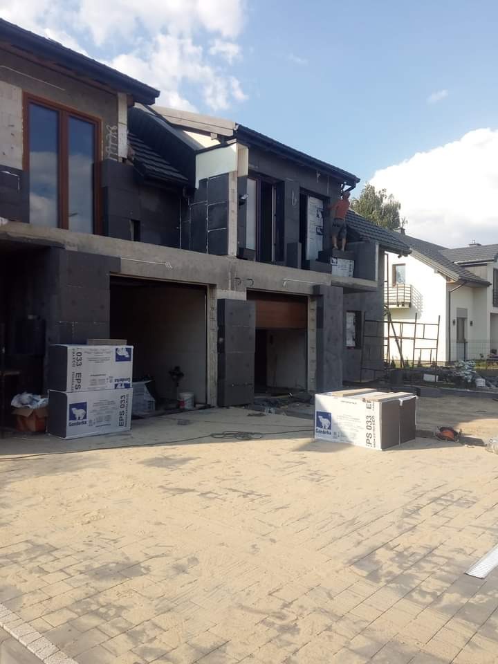 Budynek w trakcie ocieplania elewacji zewnętrznej styropianem grafitowym, widoczny pracownik na wysokości, paczki styropianu na podłożu, dom w stanie surowym otwartym.