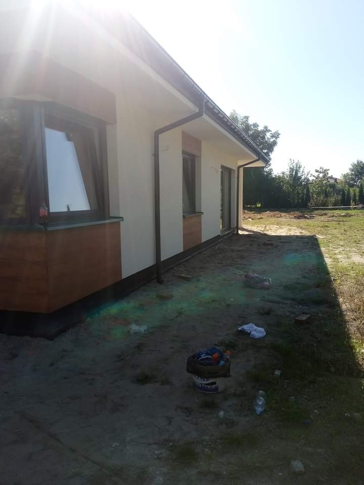 Jasna elewacja domu jednorodzinnego z brązowymi panelami pod oknami, widoczne rynny, teren wokół domu częściowo zagospodarowany, w tle zieleń drzew i krzewów, na ziemi leżą śmieci i stoi wiadro.
