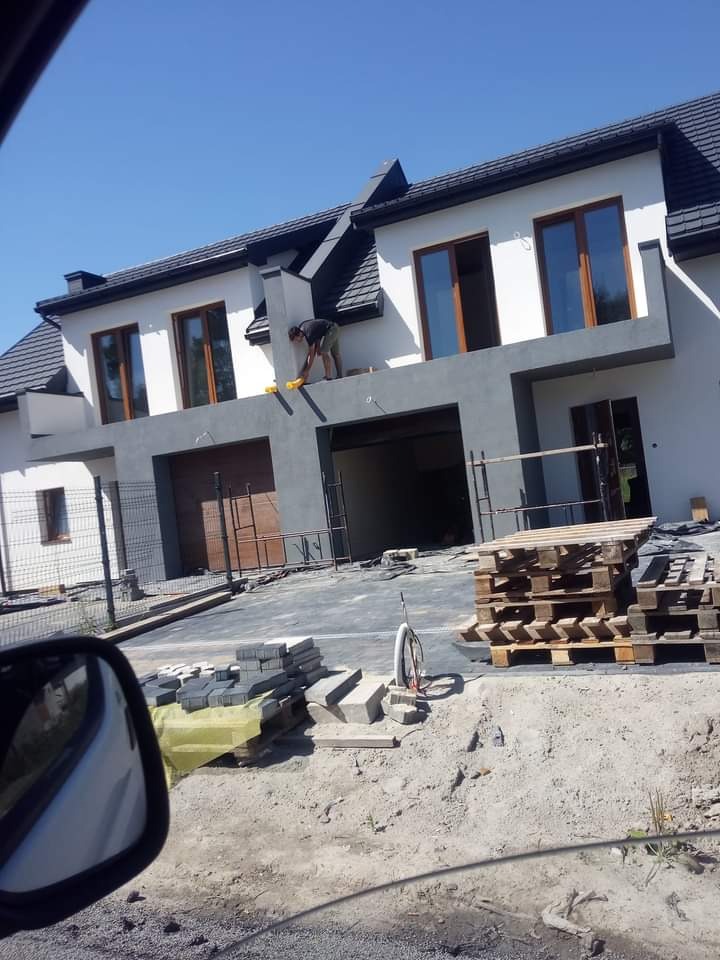 Dwukondygnacyjny dom w trakcie prac wykończeniowych elewacji, widoczny pracownik na balkonie, ułożona kostka brukowa i palety na podjeździe.