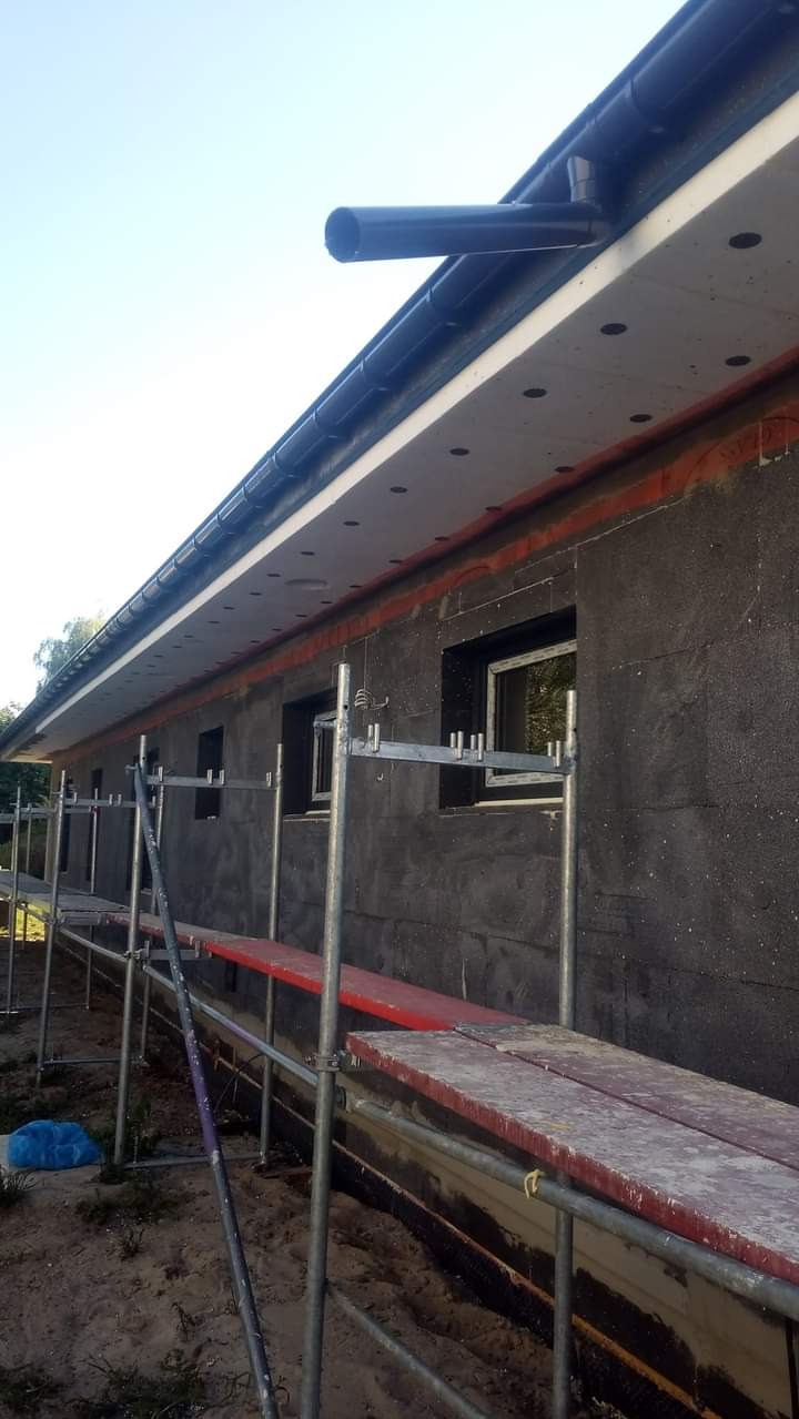 Budynek jednorodzinny w trakcie ocieplania elewacji zewnętrznej styropianem grafitowym, widoczne rusztowania i fragment orynnowania.