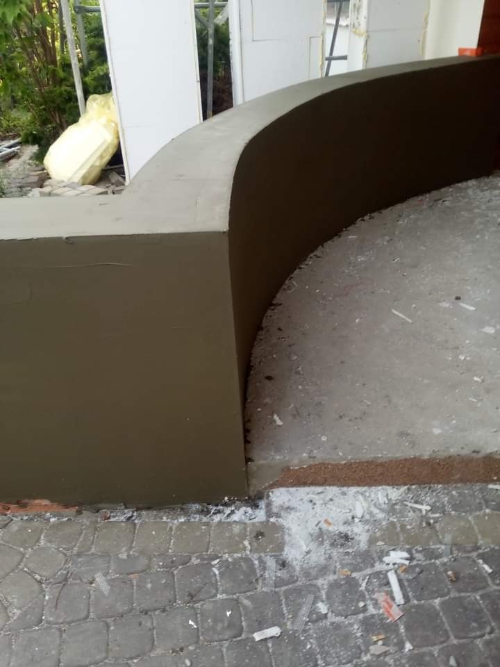 Zaokrąglona ściana elewacyjna w trakcie prac wykończeniowych, widoczne niedoskonałości tynku i fragment kostki brukowej z resztkami materiałów budowlanych.