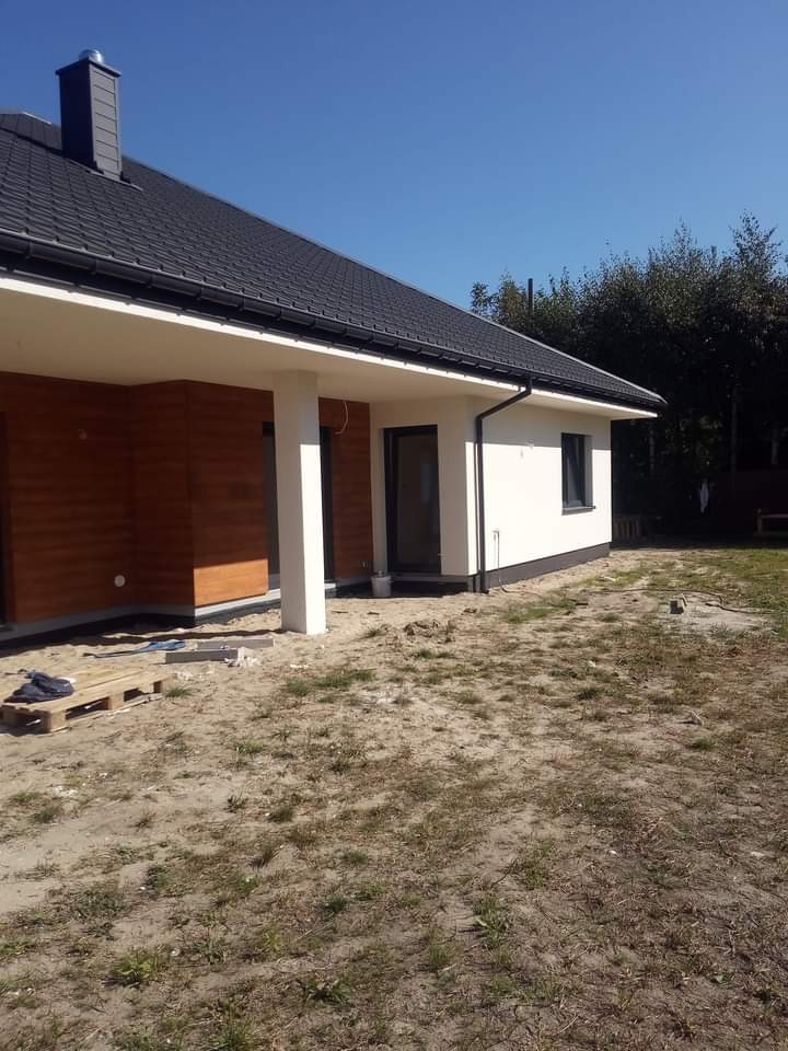 Nowoczesny dom jednorodzinny z wykończoną elewacją w kolorze białym i brązowym, widoczne elementy wykończenia, teren wokół domu częściowo zagospodarowany, paleta z materiałami budowlanymi...