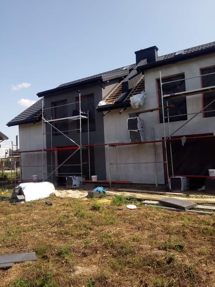Dom w trakcie prac elewacyjnych z widocznymi rusztowaniami, częściowo ocieplonym poddaszem z odkrytą konstrukcją dachu, oraz fragmentami pomalowanej i niepomalowanej elewacji.