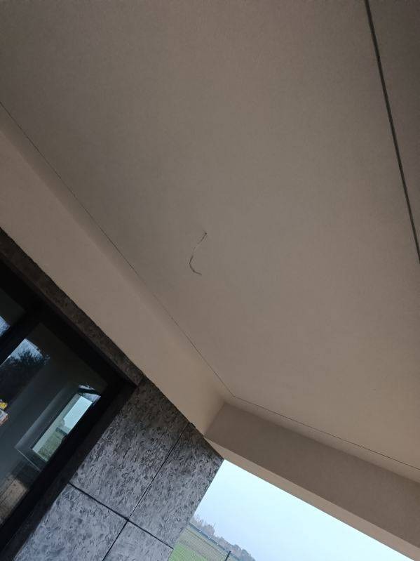 Biała, gładka elewacja podbitki dachowej z widoczną cienką rysą, fragment okna z ciemną ramą i elewacją z szarych płyt kamiennych, widok z dołu na zewnątrz budynku.