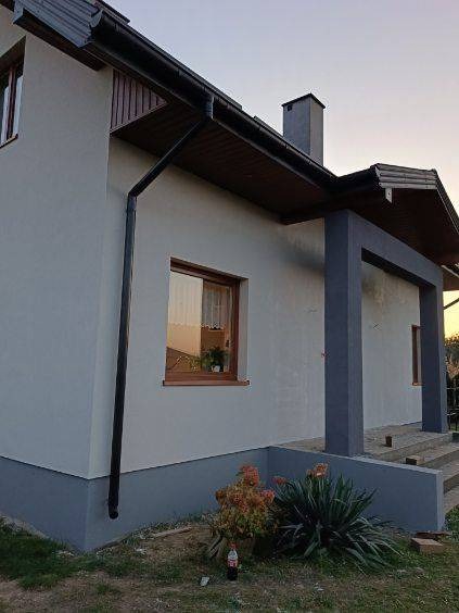 Jasnoszary dom jednorodzinny z brązowymi elementami wykończeniowymi, widoczny fragment elewacji po tynkowaniu z ciemniejszym cokołem, rynna oraz okno z kwiatami wewnątrz.