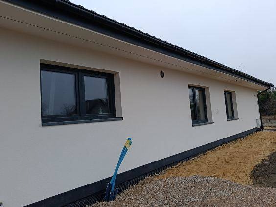 Jasna elewacja domu jednorodzinnego z grafitowymi oknami i czarną podmurówką, widok z boku na nierówny teren przed budynkiem, niebieska rura wystająca z ziemi.