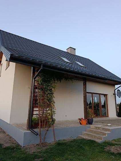 Jasny dom jednorodzinny z ciemnym dachem i oknami dachowymi, widok z boku na elewację z elementami zieleni pnącej, doniczkami i schodami.