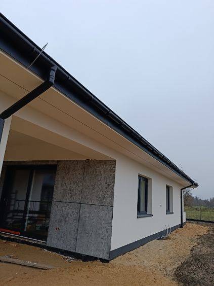 Elewacja domu jednorodzinnego w trakcie prac: fragment ściany zabezpieczony płytami OSB, świeżo pomalowana biała ściana z ciemnymi oknami i rynnami, teren wokół budynku częściowo zasypany piaskiem.