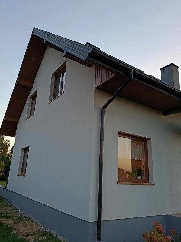 Dwupiętrowy dom z szarą elewacją i brązowymi ramami okiennymi, widoczny z boku pod lekkim kątem, z ciemnym dachem i rynnami.