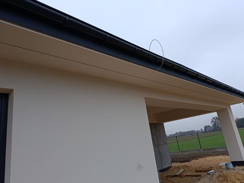 Jasna elewacja domu jednorodzinnego z czarną rynną, widoczny fragment niezakończonej obudowy filara wejścia.