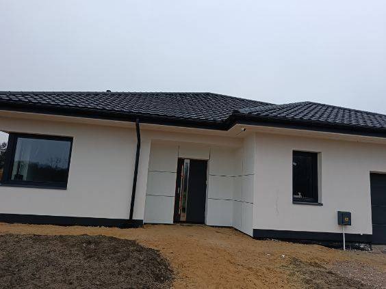 Jednopiętrowy dom z ciemną dachówką i jasną elewacją w trakcie prac wykończeniowych, widoczne fragmenty niezabezpieczonej ściany przy wejściu, teren wokół domu częściowo zagospodarowany.