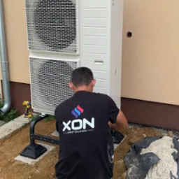 Instalacja zewnętrznej jednostki pompy ciepła Panasonic przez montera w czarnej koszulce z logo firmy XON, widoczna wiertarko-wkrętarka akumulatorowa, teren wokół częściowo wyłożony folią.