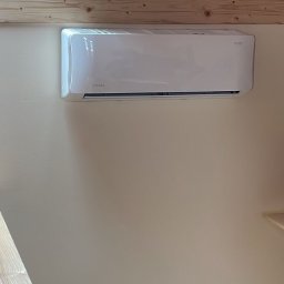 XON Łukasz Piotrowski - Biała jednostka klimatyzacyjna zamontowana na beżowej ścianie w pomieszczeniu z drewnianym stropem i widocznym gniazdkiem elektrycznym.