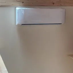 Biała jednostka klimatyzacyjna zamontowana na beżowej ścianie w pomieszczeniu z drewnianym stropem i widocznym gniazdkiem elektrycznym.