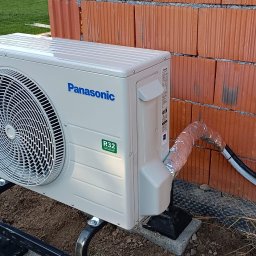 XON Łukasz Piotrowski - Zewnętrzna jednostka klimatyzacji Panasonic R32 zamontowana na czarnej metalowej podstawie przy ceglanej ścianie w trakcie instalacji, widoczne rury izolowane folią aluminiową.