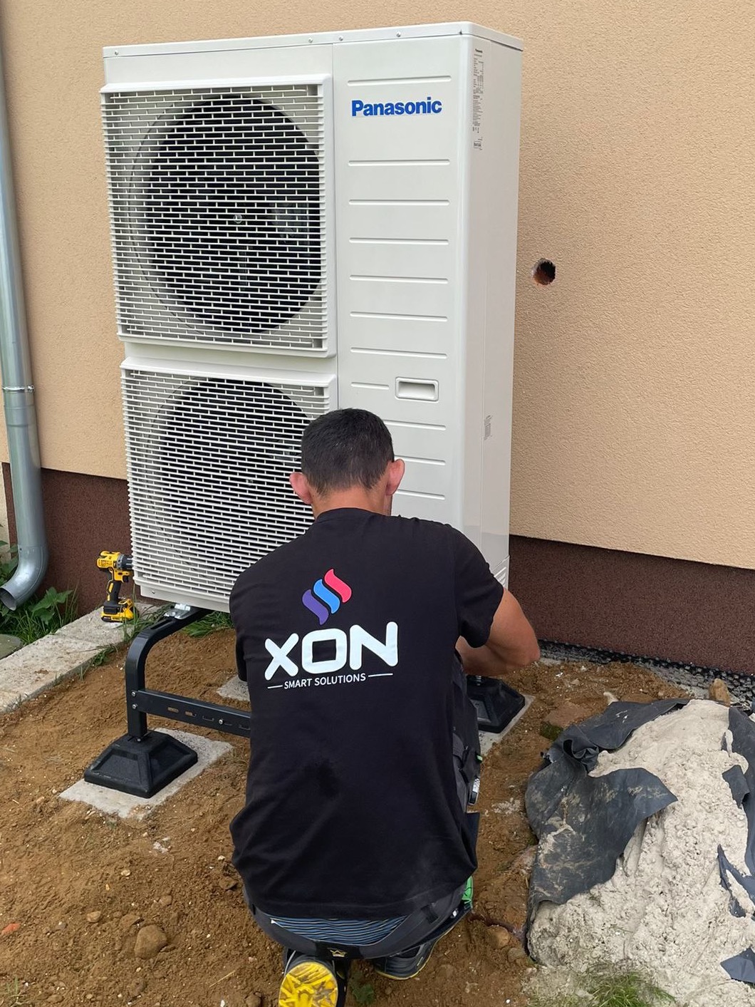 Instalacja zewnętrznej jednostki pompy ciepła Panasonic przez montera w czarnej koszulce z logo firmy XON, widoczna wiertarko-wkrętarka akumulatorowa, teren wokół częściowo wyłożony folią.