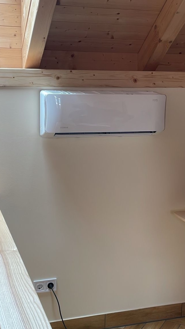 Biała jednostka klimatyzacyjna zamontowana na beżowej ścianie w pomieszczeniu z drewnianym stropem i widocznym gniazdkiem elektrycznym.