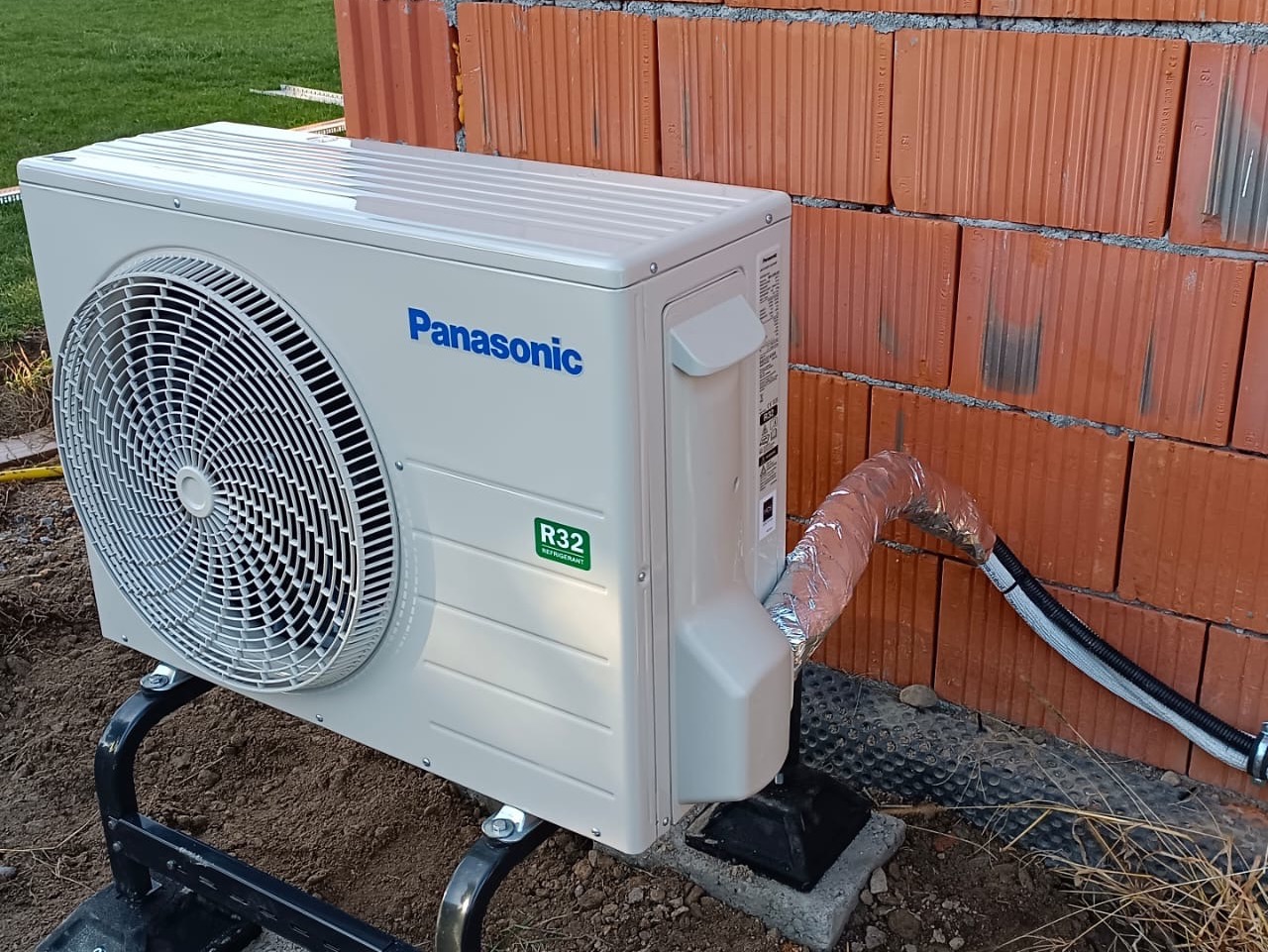 Zewnętrzna jednostka klimatyzacji Panasonic R32 zamontowana na czarnej metalowej podstawie przy ceglanej ścianie w trakcie instalacji, widoczne rury izolowane folią aluminiową.