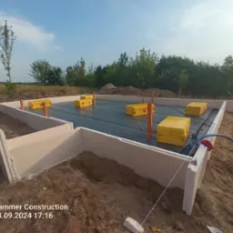 Przygotowany teren pod budowę domu: wylane fundamenty z izolacją termiczną, zabezpieczone czarną folią, widoczne żółte paczki styropianu i wystające pionowe rury.