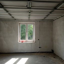 Rafal Ciekot - Wnętrze w trakcie remontu: surowe ściany z widocznymi śladami gładzi, metalowa konstrukcja podwieszanego sufitu, okno z widokiem na zieleń, wystające kable elektryczne.