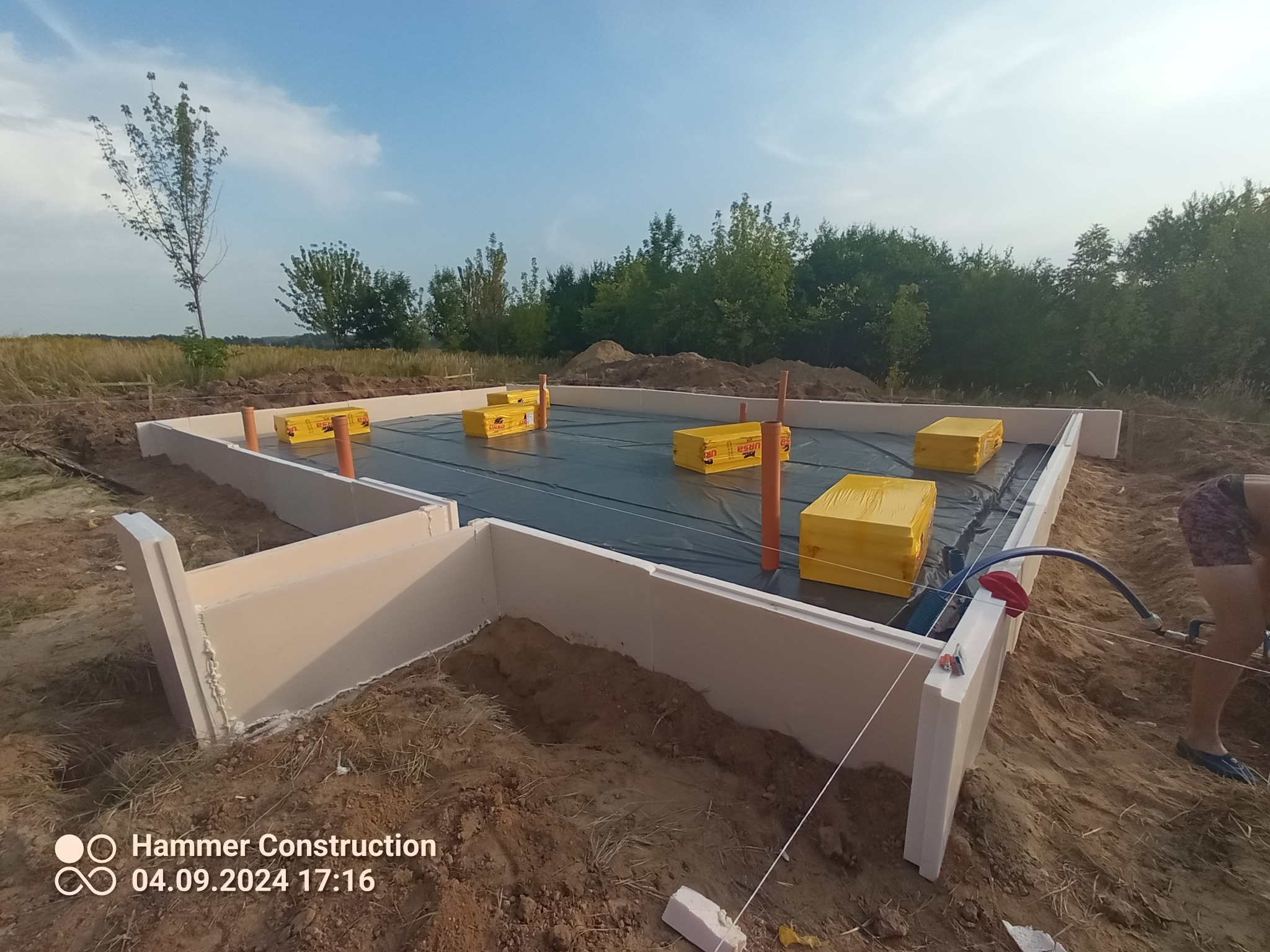 Przygotowany teren pod budowę domu: wylane fundamenty z izolacją termiczną, zabezpieczone czarną folią, widoczne żółte paczki styropianu i wystające pionowe rury.